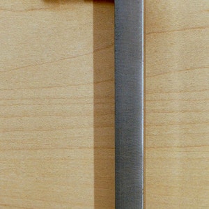 May include: A metal cabinet handle with a rectangular design. The handle is a muted silver color and is attached to a light-colored wooden surface with two screws. The handle extends vertically, creating a simple, modern aesthetic.