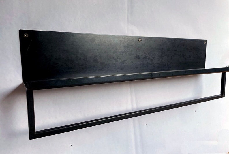 Pu&ograve; includere: Uno scaffale in metallo nero con una singola barra sottostante. Lo scaffale &egrave; montato su una parete bianca.