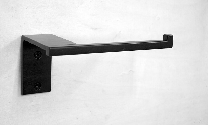 May include: Black metal toilet paper holder mounted on a white wall. The holder has a square base and a rectangular arm.