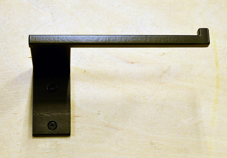 May include: Black metal toilet paper holder mounted on a wall. The holder has a rectangular base and a curved arm.