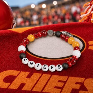 May include: A beaded bracelet featuring white, red, yellow, and black beads. The white beads spell out the word CHIEFS in black lettering. The bracelet includes a silver-toned metal charm shaped like a football and a circular metal accent piece.