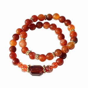 May include: Two beaded bracelets. The bracelets feature orange and red round beads with gold-colored spacers. One bracelet has a large, rectangular, dark red gem in the center. The bracelets are displayed on a white background.
