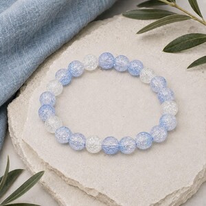 May include: A circular bracelet composed of alternating translucent light blue and clear crackle glass beads. The beads are spherical and strung together in a repeating pattern, creating a textured appearance with a reflective surface finish.