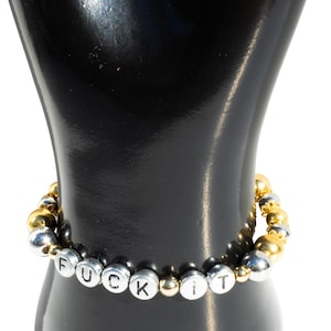 May include: A beaded bracelet with silver and gold-colored beads. The bracelet has the phrase "FUCK IT" spelled out in silver letter beads. The bracelet is displayed on a black mannequin hand.