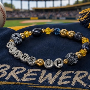 May include: A beaded bracelet featuring silver-toned letter beads spelling CLAWS UP, interspersed with black crackle-patterned beads, metallic gold spheres, faceted yellow crystals, and a single dark blue bead, arranged in a circular pattern.