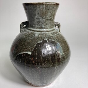 May include: A handcrafted ceramic vase with a dark, mottled glaze. The vase features a rounded body, a flared neck, and two small handles. The color palette includes shades of brown, green, and white, creating a rustic and organic aesthetic.