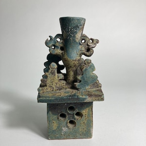 Puede incluir: Escultura de cerámica artesanal en tonos turquesa y marrón. Presenta una base cuadrada con cuatro aberturas circulares, una plataforma rectangular y una estructura central en forma de jarrón con elementos decorativos.