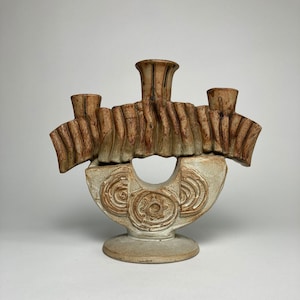 Puede incluir: Un candelabro de cerámica con un diseño único. El portavelas presenta tres copas para velas y una copa central, todas en un color marrón cálido. La base es de color beige claro con patrones circulares tallados. La forma general es abstracta y artística.