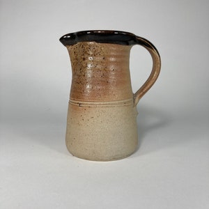 May include: A handmade ceramic jug with a dark brown rim and handle. The body transitions from a light beige base to a speckled, tan middle section, and a darker brown top. The jug has a curved handle and a spout for pouring.