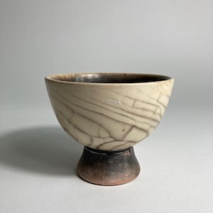 Può includere: Una ciotola in ceramica con esterno color crema e interno marrone scuro. La ciotola ha una base a piedistallo e un motivo screpolato a forma di ragnatela all'esterno. Il bordo è marrone scuro e la ciotola è posta su uno sfondo neutro.