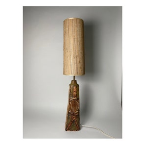 Puede incluir: Una lámpara de cerámica vintage alta con una pantalla cilíndrica texturizada. La base de la lámpara es de color verde oscuro y marrón con diseños geométricos tallados. La pantalla es de color beige claro.