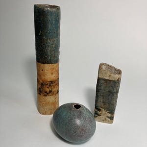 Puede incluir: Colección de jarrones de cerámica hechos a mano y un jarrón pequeño y redondeado. El jarrón alto es cilíndrico con una combinación de colores azul y beige. El jarrón pequeño tiene forma ovalada con un esmalte turquesa. El jarrón rectangular tiene una superficie texturizada.