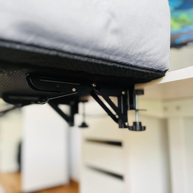May include: A black, adjustable desk arm with a clamp-on design. The arm is attached to a white desk and supports a gray, cushioned surface. The clamp features a black knob for tightening and adjusting the arm's position.