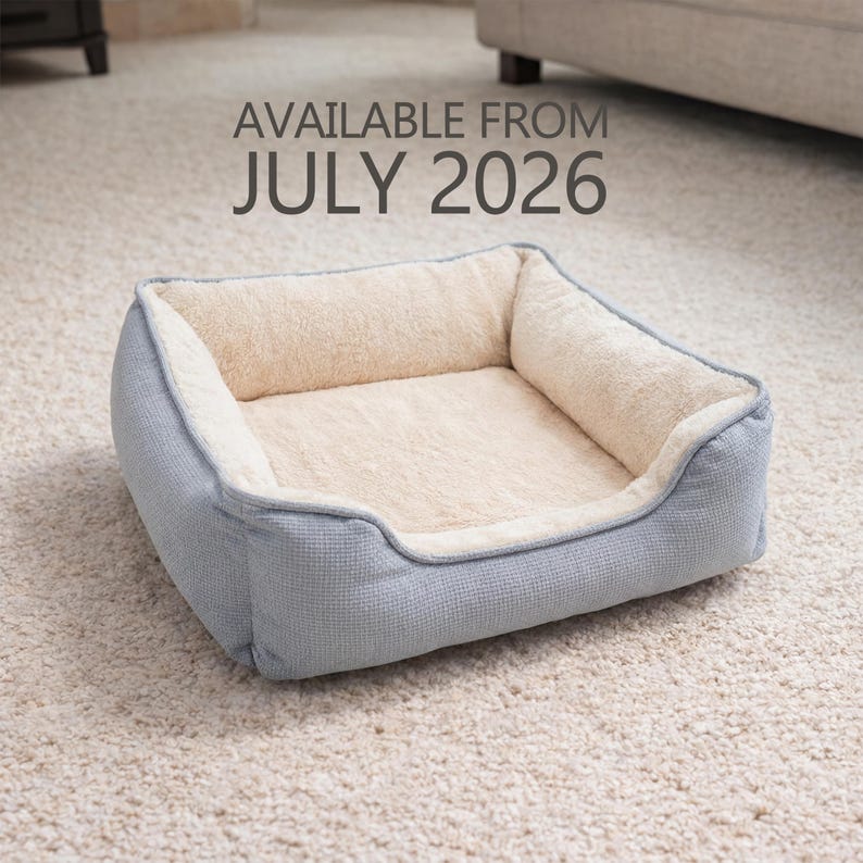 May include: A light blue pet bed with a soft, cream-colored interior. The bed has a rectangular shape with raised sides and a plush, cushioned surface. The text "AVAILABLE FROM JULY 2026" is displayed above the bed.