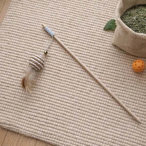 Könnte beinhalten: Ein Katzenspielzeug mit einem langen Holzstab, einem gewebten Ball, einer Feder und einer goldenen Perle. Ein kleiner orangefarbener Ball und ein Jutesack mit grünem Katzenminze sind ebenfalls zu sehen.