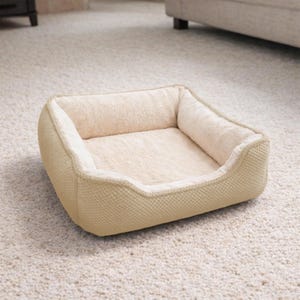 May include: A beige dog bed with a soft, plush interior and a textured exterior. The bed has a rectangular shape with raised sides for comfort and security. The bed is placed on a light-colored carpet.