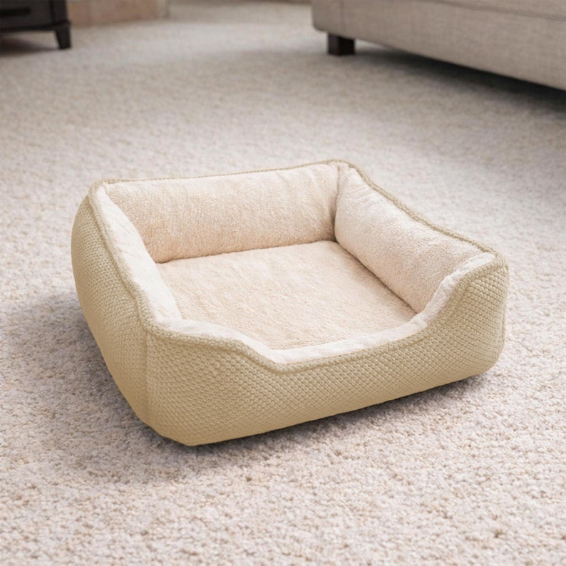 May include: A beige dog bed with a soft, plush interior and a textured exterior. The bed has a rectangular shape with raised sides for comfort and security. The bed is placed on a light-colored carpet.