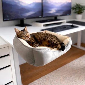 Puede incluir: Un gato atigrado marrón duerme plácidamente en una cama para mascotas gris y crema, sujeta al borde de un escritorio blanco. La cama para gatos está suspendida y el gato es el foco principal. El fondo incluye monitores de ordenador y un teclado.