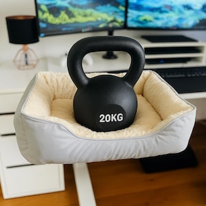 May include: A black kettlebell, labeled "20KG", rests inside a light gray pet bed with a cream-colored, fluffy interior. The bed has a rectangular shape and is placed on a white surface. The background includes a desk and computer monitor.