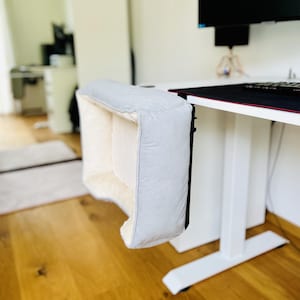 May include: A light gray pet bed, designed to attach to a desk, with a soft, cream-colored interior. The bed is rectangular and has a padded exterior. The desk is white, and the floor is hardwood.