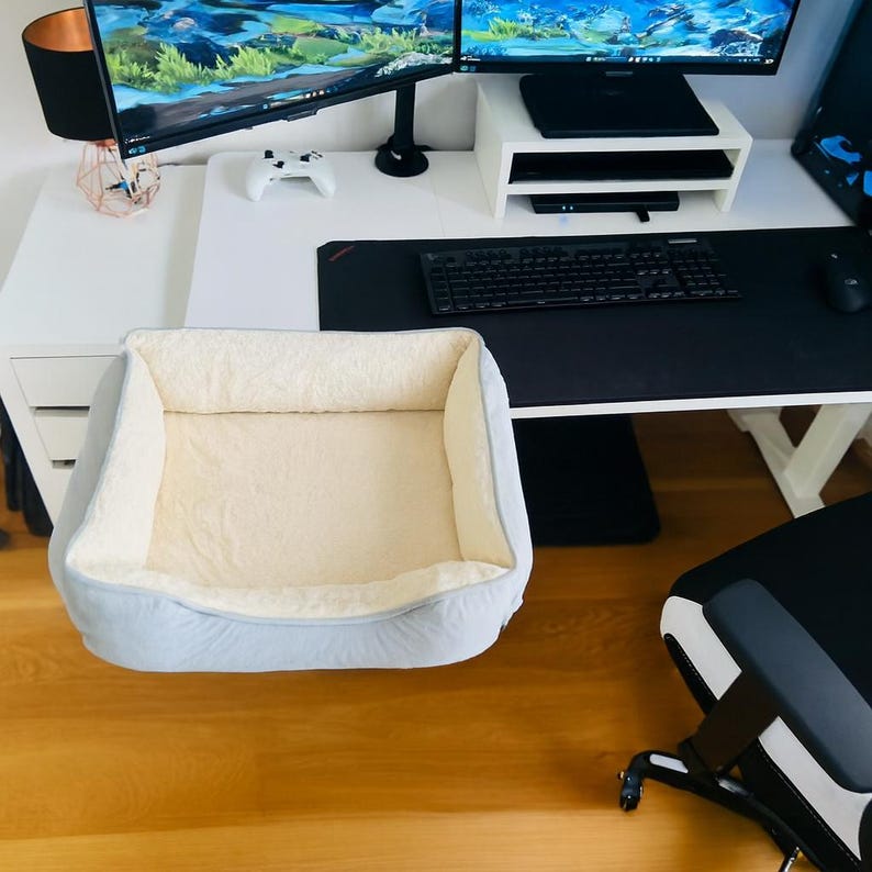 May include: A light blue pet bed with a cream-colored, plush interior sits on a wooden floor. The bed is rectangular with a soft, raised edge. The background includes a white desk, computer monitors, and a black keyboard.