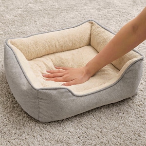May include: A rectangular pet bed with a gray exterior and a plush, cream-colored interior. The bed has a soft, cushioned appearance, and a hand is resting on the interior, suggesting a comfortable texture. The bed is designed for small pets.