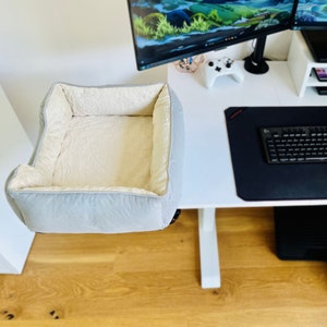 May include: A light gray pet bed with a plush, cream-colored interior. The bed is rectangular with raised sides and is designed to be placed on a desk or similar surface. The bed is made of soft, comfortable materials.