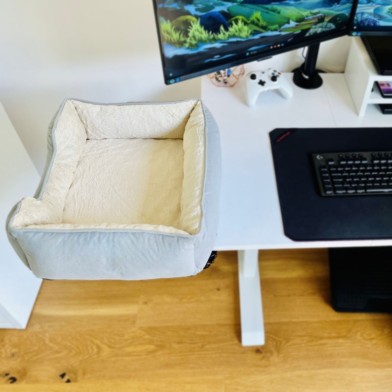 May include: A light gray pet bed with a plush, cream-colored interior. The bed is rectangular with raised sides and is designed to be placed on a desk or similar surface. The bed is made of soft, comfortable materials.