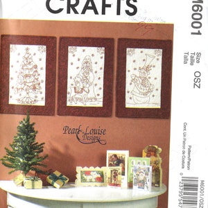 May include: A McCall's Crafts pattern featuring three framed Christmas designs: a tree, Santa with a reindeer, and a snowman. The image also shows a small Christmas tree, wrapped gifts, and greeting cards on a white table.