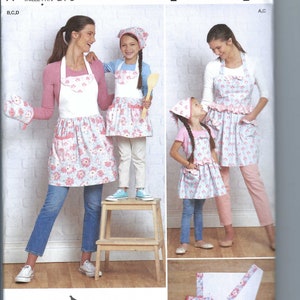 May include: A sewing pattern for a set of three aprons with a floral print. The pattern includes a large apron, a medium apron, and a small apron. The aprons are all white with a pink and blue floral print. The pattern also includes a set of matching oven mitts and a tea towel.