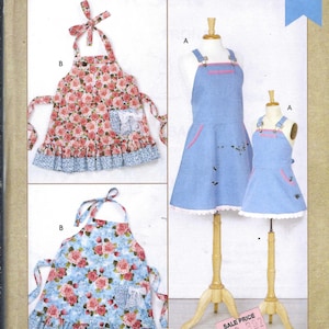 May include: Kwik Sew pattern for children's and misses' aprons. The image shows aprons with floral and solid designs. One apron has a pink and white floral pattern, while another has a blue floral pattern. The aprons are displayed on mannequins.