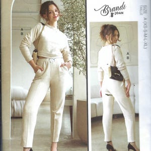 May include: McCall's M8447 sewing pattern envelope featuring Brandi Joan designs. The front shows a cream-colored long-sleeve cropped sweatshirt and matching high-waisted trousers with a drawstring waist. Includes text M8447, Size A, and a QR code.