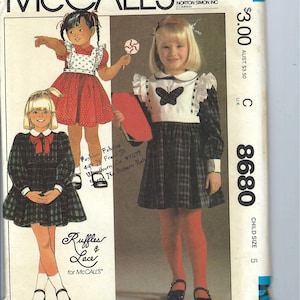 May include: A vintage McCall's sewing pattern for a child's dress. The pattern is for size 5 and features two different styles of dresses. One dress is red with white polka dots and the other is black and white plaid. The pattern includes instructions for making both dresses.
