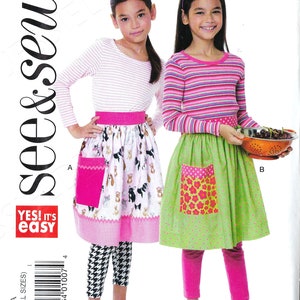 May include: A sewing pattern for two children's aprons. The pattern includes two different styles, one with a pink and white dog print and the other with a green and pink floral print. The pattern is from the brand "see & sew" and is labeled "B6611".