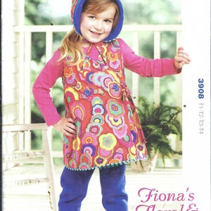 May include: A child wearing a red floral tunic, pink long-sleeve shirt, blue pants, and a blue and floral hood. The tunic has a colorful floral pattern. The image is from a Kwik Sew pattern, labeled "Fiona's Floral & Fleece."