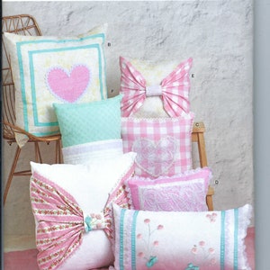 May include: A sewing pattern for making five different cushion designs. The cushions are all in shades of pink, white, and blue with various patterns and embellishments. The pattern is called Simplicity S9574 and is for one size.