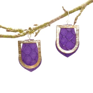 Peut inclure: Une paire de boucles d'oreilles en cuir violet avec des montures en métal doré. Les boucles d'oreilles ont la forme d'un bouclier et une surface texturée.