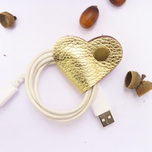 May include: A gold heart-shaped leather cord organizer with a brass button. The organizer is holding a white cord.