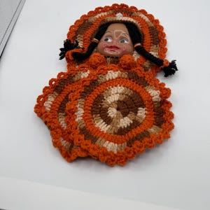 Op de afbeelding: Een vintage pop met een geschilderd gezicht en zwarte vlechten, gehuld in een handgemaakt gehaakt outfit. De outfit heeft concentrische cirkels van oranje, bruin en beige garen, met een geschulpte rand. De hoed van de pop past bij het kleurenschema van de outfit.