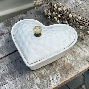 Op de afbeelding: Een witte hartvormige keramische schaal met een textuuroppervlak en een doorzichtige glazen knop. De schaal staat op een houten oppervlak, met gedroogde bloemen op de achtergrond. De schaal is waarschijnlijk voor serveren of opslag.