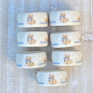 May include: Seven white ceramic napkin rings with a light blue border and a repeating pattern of teddy bears and small floral designs. Each ring features a single teddy bear with a blue bow.