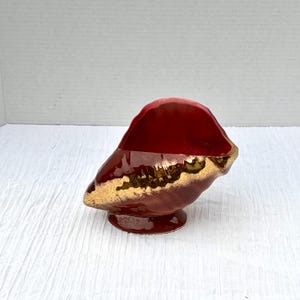 Może przedstawiać: Ceramiczny element dekoracyjny z błyszczącą czerwoną górą i złotą obwódką. Podstawa ma pasujący czerwony kolor. Element ma unikalny, organiczny kształt i spoczywa na małej, okrągłej podstawie.