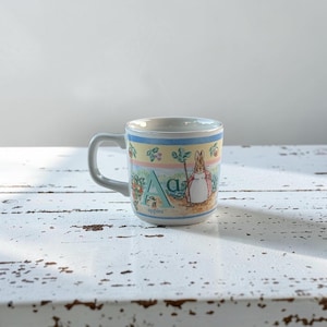 May include: A white ceramic mug featuring a colorful illustration of a rabbit character, the letter A, and the word apples. The design includes a pastel-colored border with small fruit motifs, a handle on the left side, and a smooth, glossy finish.
