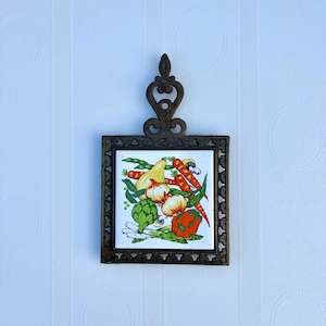 May include: A vintage metal trivet with a square ceramic tile featuring a colorful illustration of vegetables. The vegetables include carrots, onions, a pear, a red bell pepper, and artichokes. The trivet has a decorative handle.