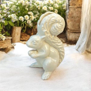 May include: A ceramic squirrel figurine in a soft, off-white color. The squirrel is depicted in a seated position, holding a nut, with a large, detailed tail. The figurine has a glossy finish and is likely a decorative item for a home.