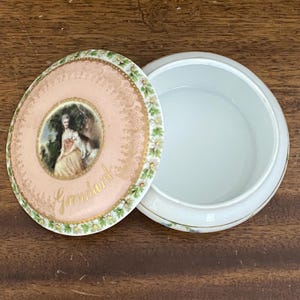 Puede incluir: Una caja de baratijas redonda de porcelana, con la tapa abierta. La tapa es de color rosa pálido con un borde dorado y una pintura de una mujer y un perro. El interior de la caja es blanco y el borde tiene un ribete dorado. La tapa está decorada con detalles florales y dorados.