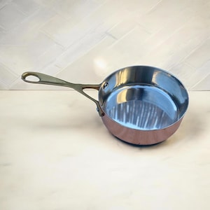 Pode incluir: Uma panela de cor cobre com interior em aço inoxidável e uma longa alça dourada. A panela é redonda com uma borda ligeiramente alargada. A alça tem um orifício para pendurar. A panela está vazia e está sobre uma superfície clara.