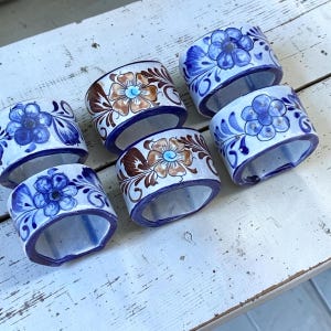 Puede incluir: Juego de seis servilleteros de cerámica. Cada anillo presenta un diseño floral pintado a mano en azul, marrón y blanco, con un borde azul. Los anillos están dispuestos sobre una superficie de madera blanca envejecida. Ideales para una mesa elegante.