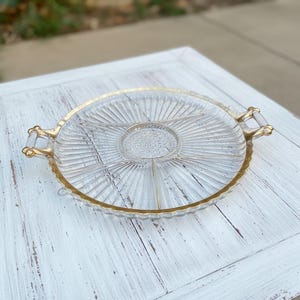 Puede incluir: Una bandeja de servir de vidrio transparente con borde y asas doradas. La bandeja está dividida en secciones y presenta un diseño radial. Las asas son doradas y transparentes. La bandeja está sobre una superficie blanca.