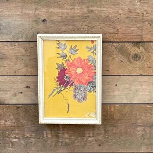 May include: A framed floral print featuring a bouquet of flowers in shades of coral, burgundy, and gray against a yellow background. The print is in a white ornate frame, creating a vintage aesthetic. The artwork is displayed on a wooden surface.